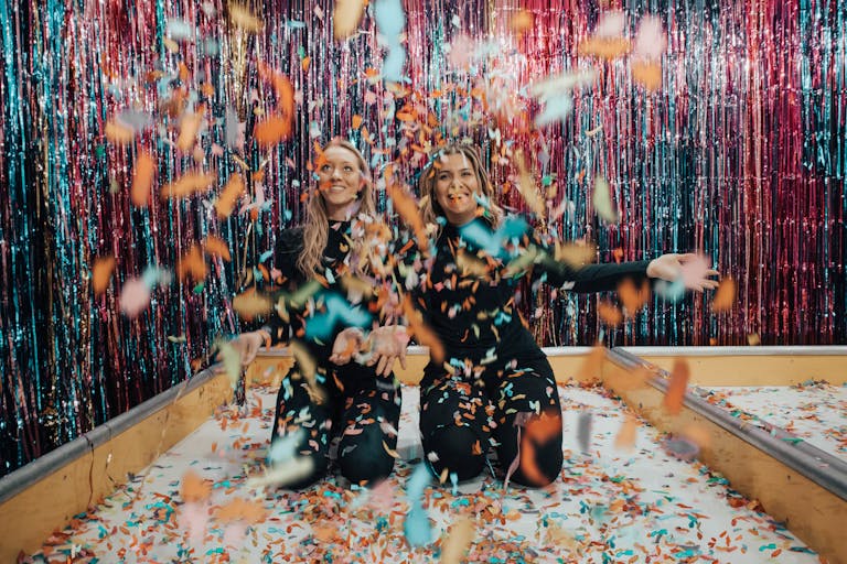Two women enjoying a vibrant indoor celebration with colorful confetti.